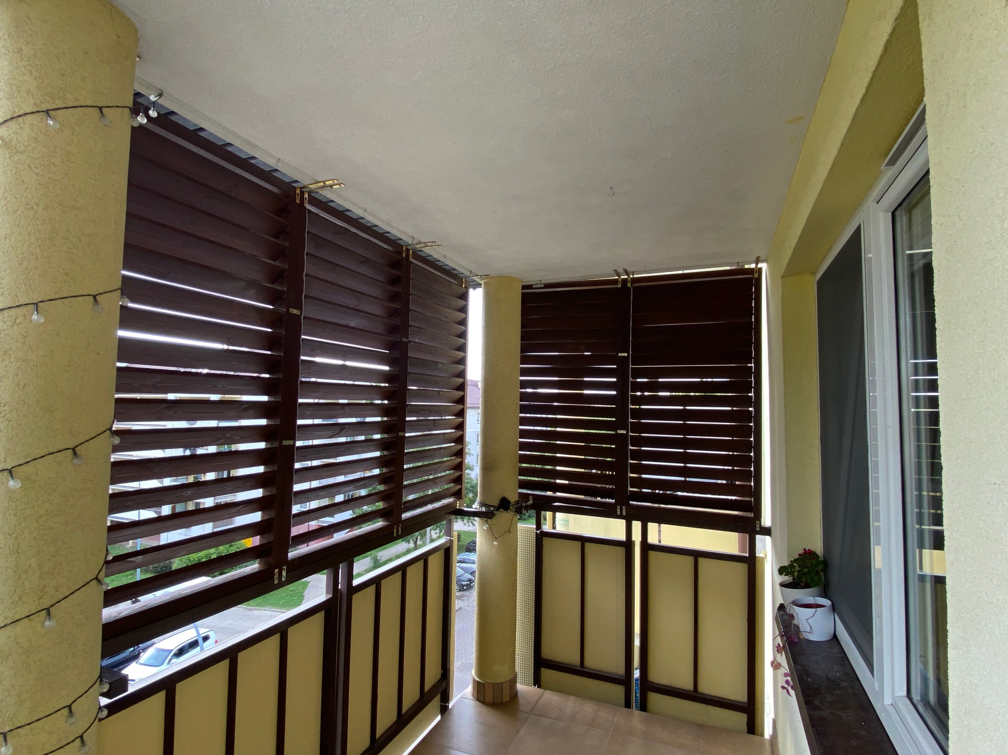 Zadaszony balkon z drewnianymi żaluzjami w kolorze ciemnego brązu, dekorowany girlandą świetlną, widok na ulicę i budynki w tle.