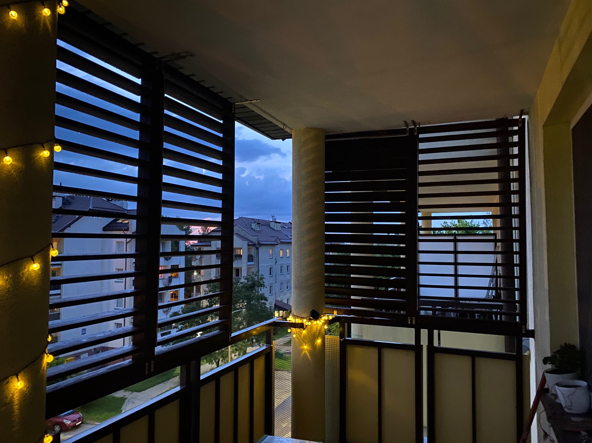 Zadaszony balkon z drewnianymi żaluzjami, oświetlony girlandą z ciepłych światełek, z widokiem na budynki mieszkalne i zachodzące słońce.