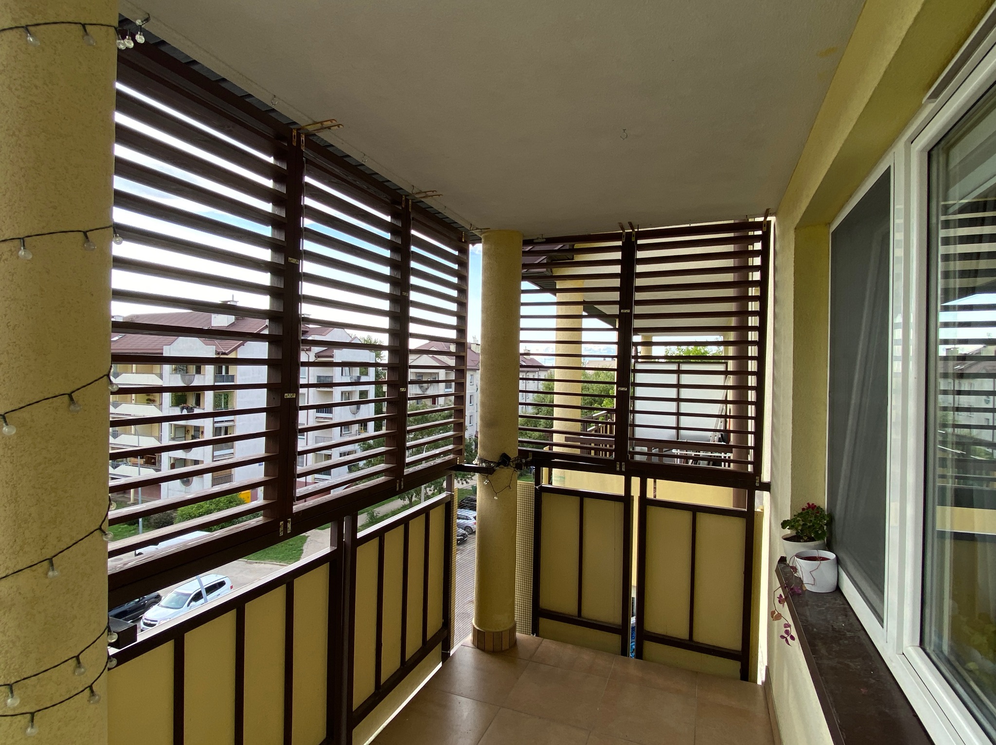 Zadaszony balkon z drewnianymi żaluzjami i balustradą, oświetlony girlandą z żarówek, z widokiem na budynki mieszkalne.