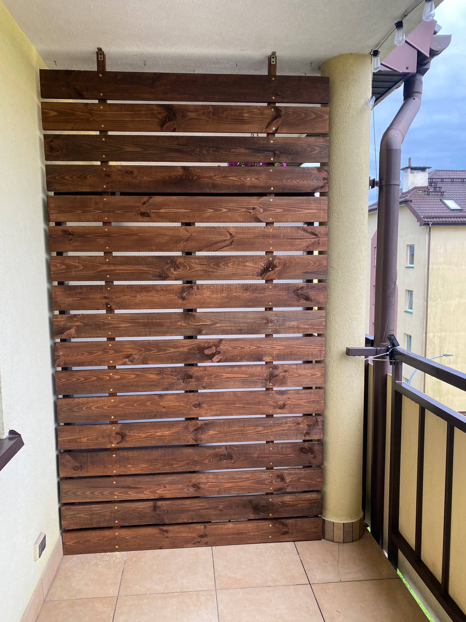 Panel z ciemnego drewna na balkonie, poziome deski z widocznymi śrubami, zamontowany między ścianą a kolumną, z widokiem na sąsiednie budynki.