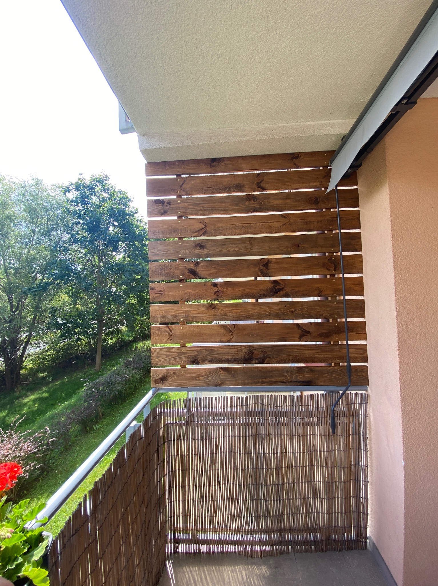 Balkon z drewnianą osłoną z desek na górze i bambusową na dole, częściowo zasłonięta markiza, widok na zieleń za balkonem.