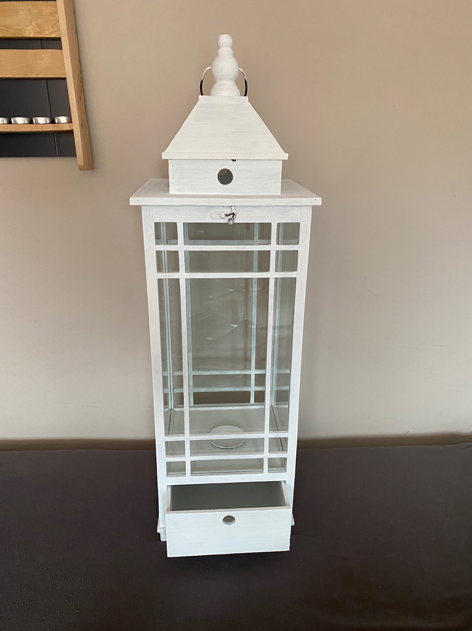 Biała, drewniana latarnia dekoracyjna z szybkami i wysuwaną szufladką, stojąca na ciemnym blacie, na tle beżowej ściany z drewnianą półką na tealighty po lewej stronie.