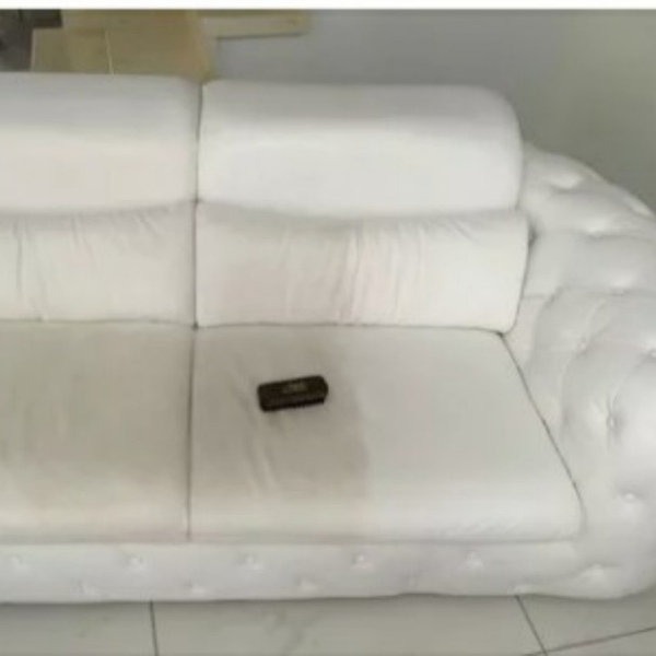 Biała, skórzana kanapa z widoczną różnicą w czystości pomiędzy lewą i prawą poduszką siedziska; na prawej poduszce leży ciemne pudełko.