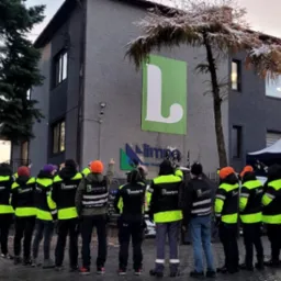 Grupa pracowników w odblaskowych kurtkach firmy Lembro, stojących przed budynkiem z logo firmy Limpa i wskazujących na coś na dachu budynku.