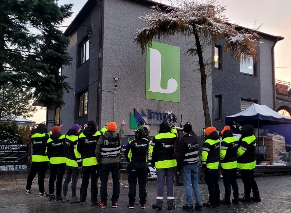 Grupa pracowników w odblaskowych kurtkach firmy Lembro, stojących przed budynkiem z logo firmy Limpa i wskazujących na coś na dachu budynku.