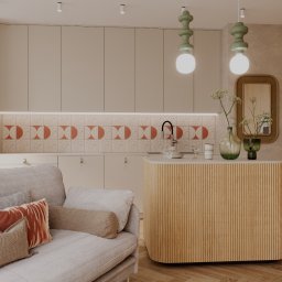 Archiidea Piotr Sekulski - Beżowa kuchnia z wyspą, terakotowymi płytkami i drewnianymi szafkami, widok z salonu z szarą sofą i poduszkami w odcieniach brązu i pomarańczy.