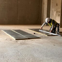 Karpińscy wentylację Kamil Karpiński - Pracownik w kasku i kamizelce odblaskowej, klęcząc na betonowej podłodze, mierzy i przycina nożem płytę izolacyjną z folią aluminiową, rozłożoną na podłodze w surowym wnętrzu budynku.