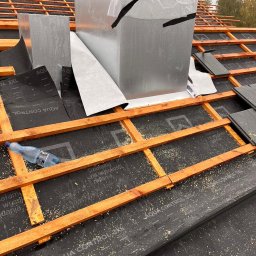 Karpińscy wentylację Kamil Karpiński - Montaż blaszanej obudowy komina na dachu z drewnianą konstrukcją, widoczna czarna membrana dachowa i częściowo ułożone dachówki, pracownik w odblaskowej kamizelce przytrzymuje element konstrukcji.