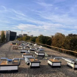 Widok z góry na liczne moduły wentylacyjne na dachu pokrytym żwirem, w tle budynki miejskie i niebieskie niebo z chmurami, mała postać ludzka w odblaskowej kamizelce przy jednym z modułów.