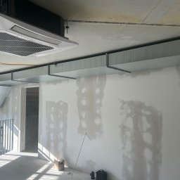 Karpińscy wentylację Kamil Karpiński - Wnętrze w trakcie wykańczania z zamontowaną kasetą klimatyzacyjną sufitową i kanałami wentylacyjnymi pod stropem. Ściany z widocznymi śladami szpachlowania, podłoga z resztkami gruzu.