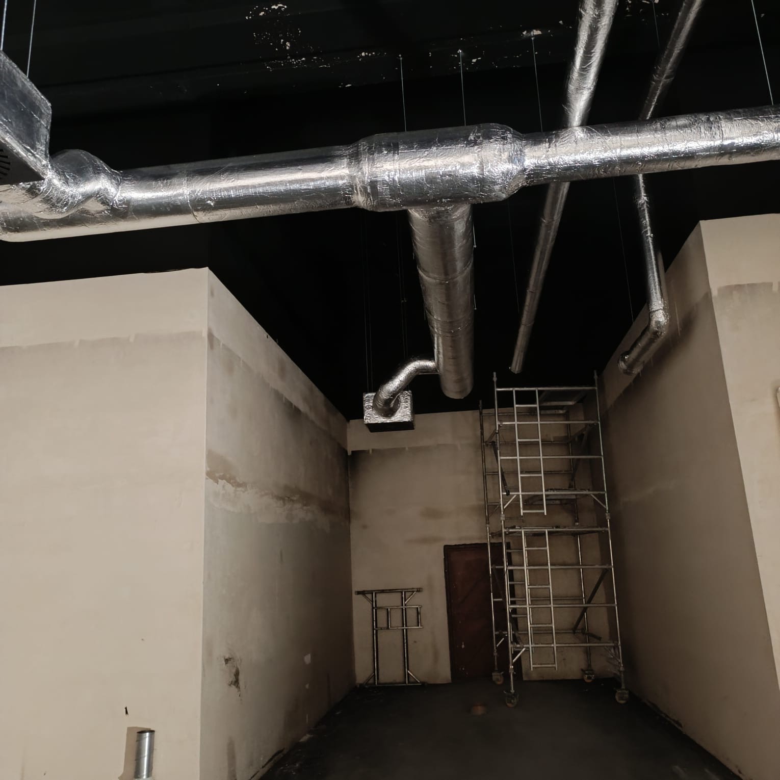 Wnętrze w trakcie instalacji systemu wentylacyjnego z widocznymi izolowanymi rurami pod sufitem, rusztowaniem i surowymi ścianami.