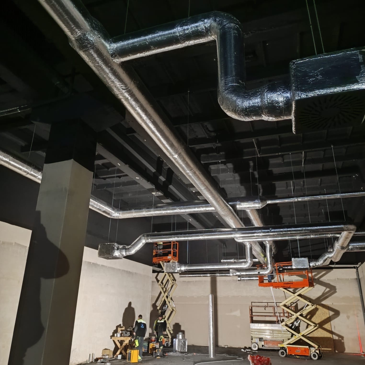 Montaż izolowanych przewodów wentylacyjnych pod sufitem w hali, widoczne podnośniki nożycowe i sylwetki pracowników.