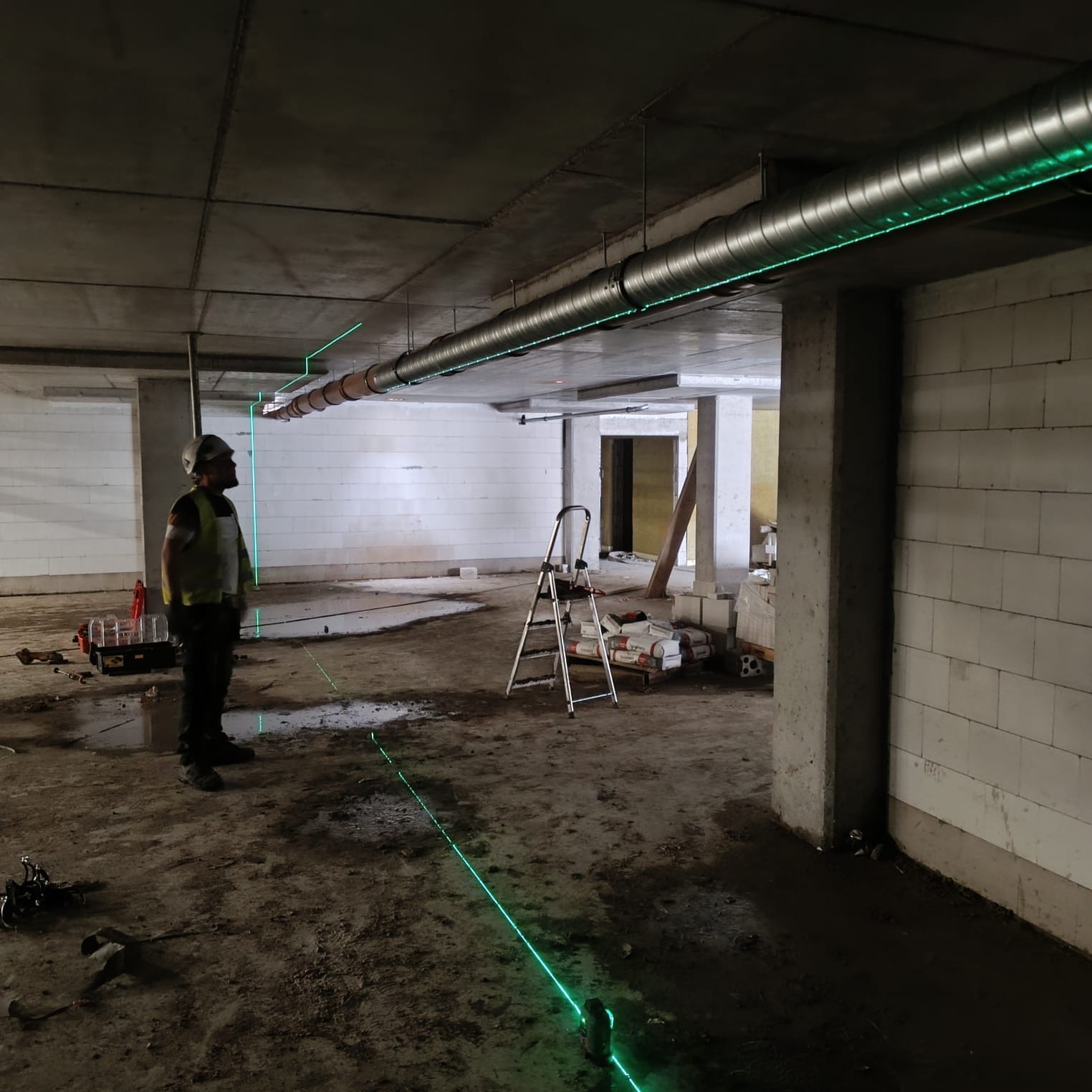 Instalacja wentylacji: pracownik w kasku i kamizelce odblaskowej kontroluje poziom rur wentylacyjnych pod sufitem za pomocą lasera krzyżowego w surowym wnętrzu budynku w trakcie budowy.