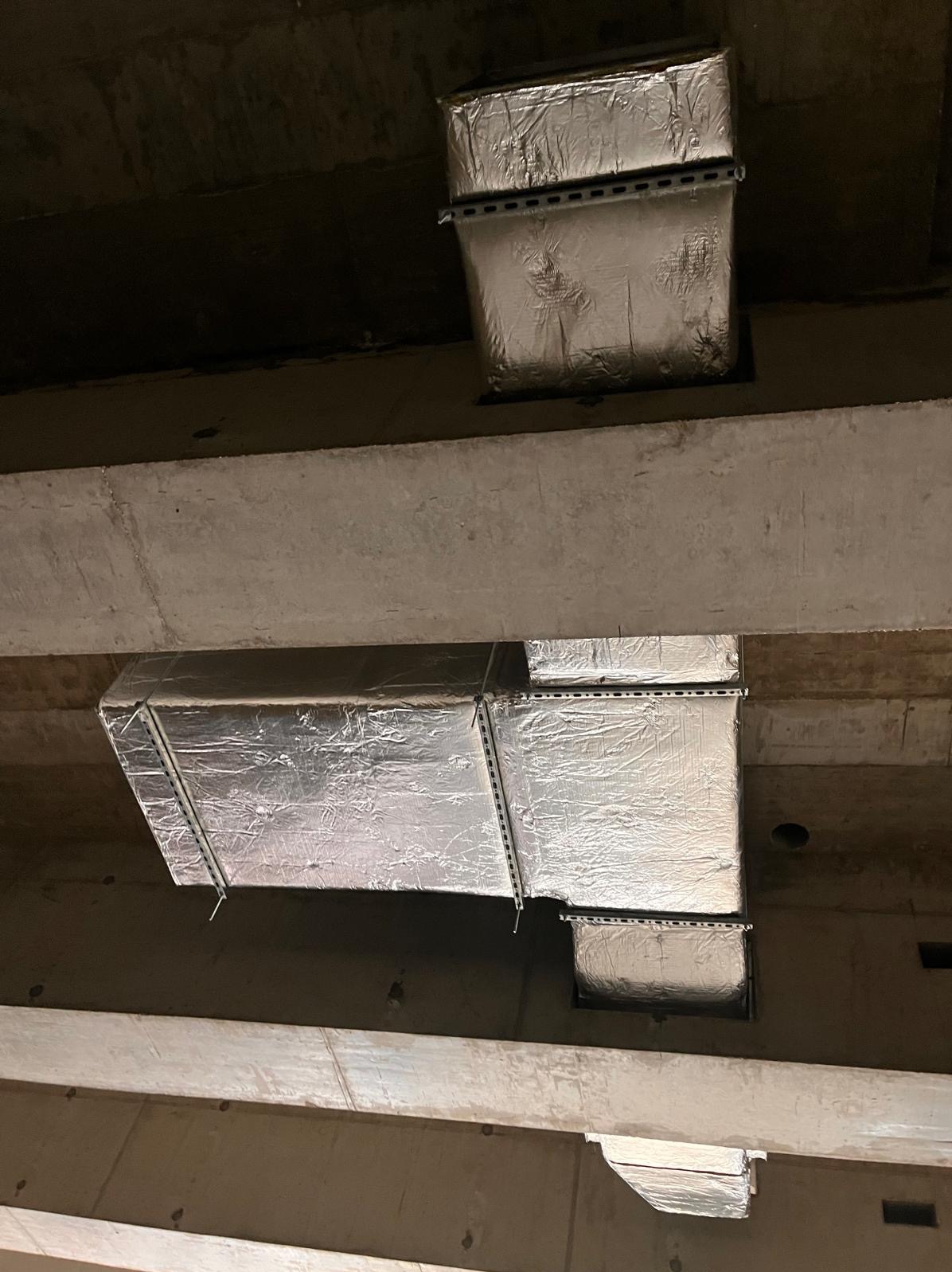 Izolowane kanały wentylacyjne zawieszone pod betonowym stropem, wsparte na perforowanych profilach montażowych.