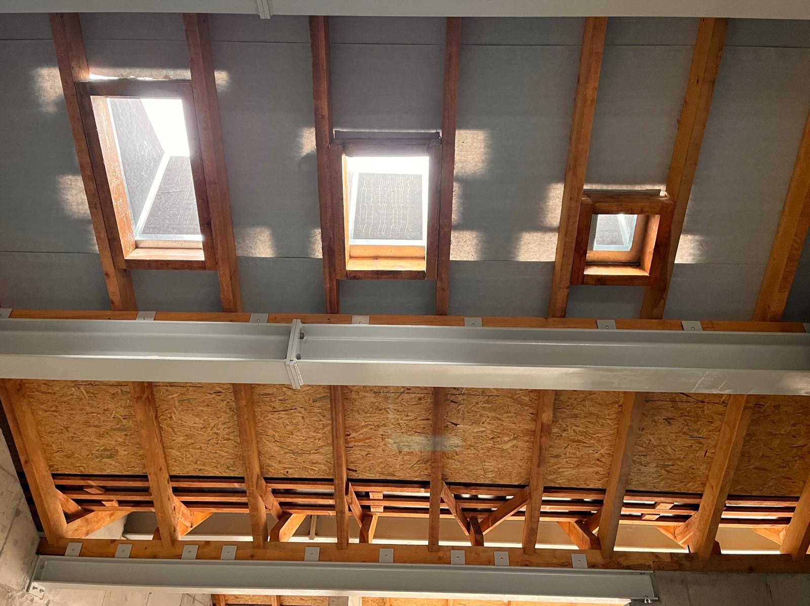 Widok z dołu na drewnianą konstrukcję dachu z trzema oknami dachowymi o różnej wielkości, z widoczną szarą membraną dachową i stalową belką nośną.