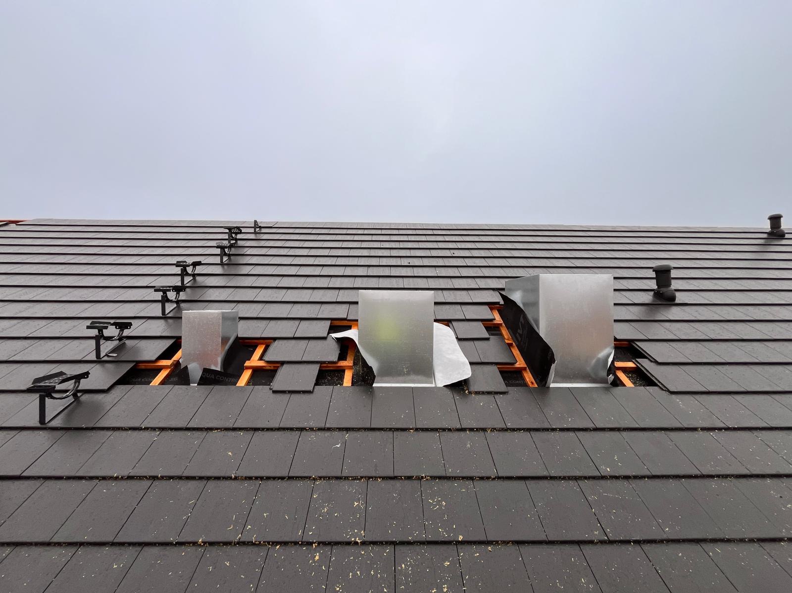 Montaż systemów wentylacyjnych na dachu z ciemnej dachówki, widoczne metalowe kanały wentylacyjne i elementy konstrukcyjne mocowań.