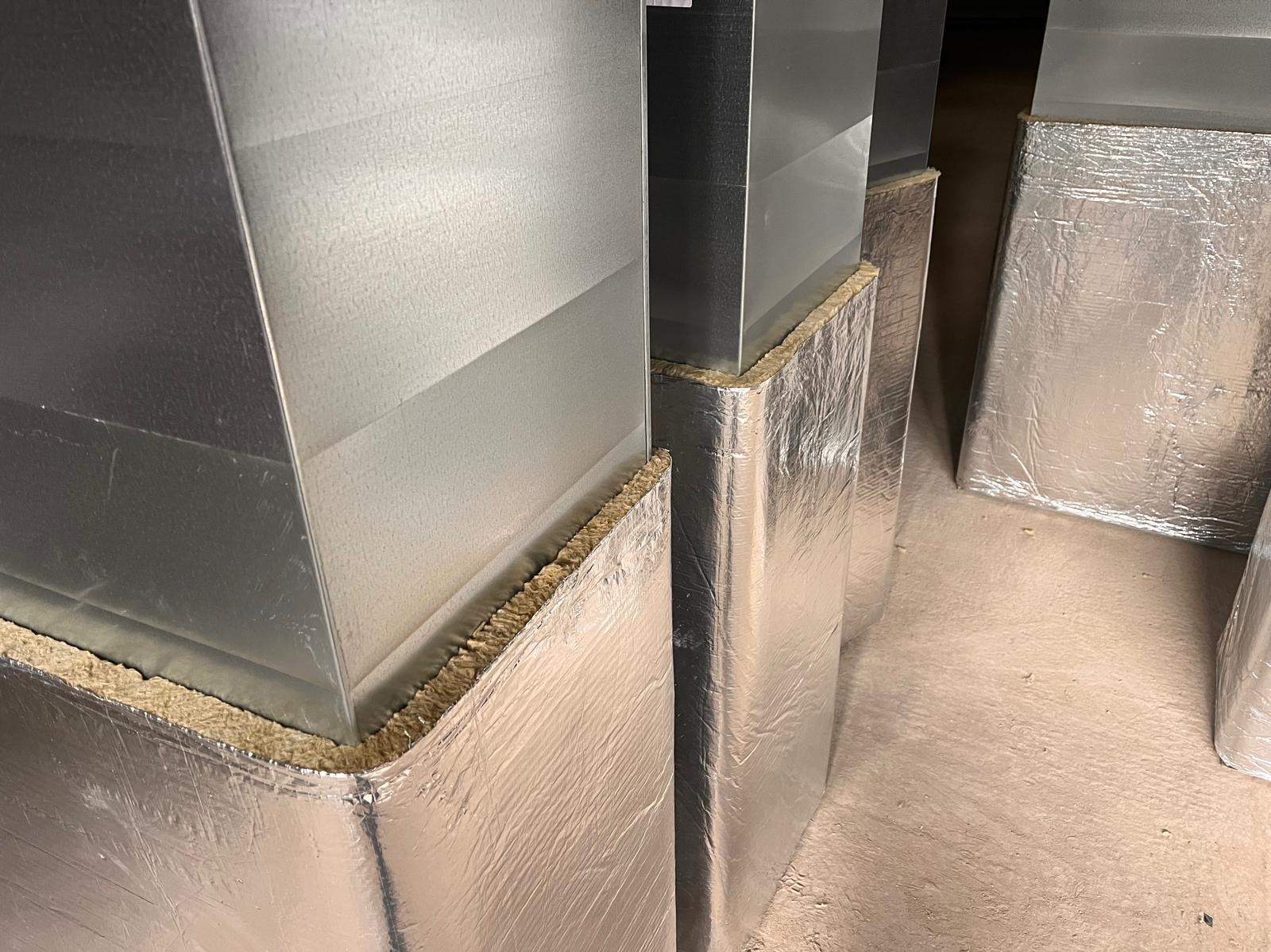 Metalowe kanały wentylacyjne z izolacją z wełny mineralnej i folii aluminiowej, widoczne łączenia i struktura materiałów.