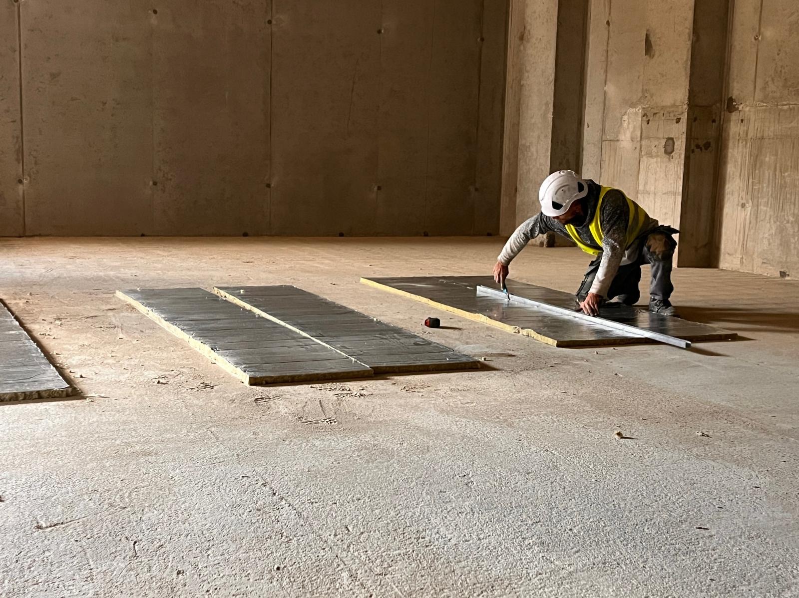 Pracownik w kasku i kamizelce odblaskowej, klęcząc na betonowej podłodze, mierzy i przycina nożem płytę izolacyjną z folią aluminiową, rozłożoną na podłodze w surowym wnętrzu budynku.