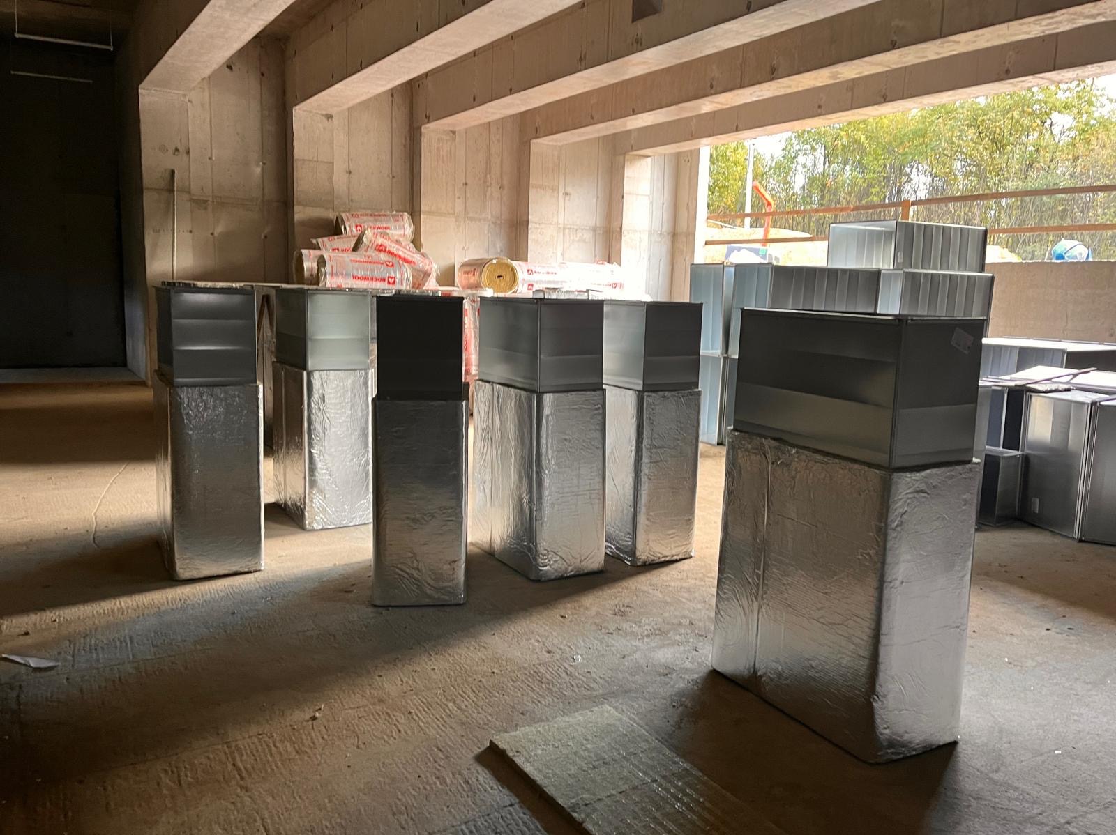 Przygotowane do montażu, zaizolowane elementy systemu wentylacyjnego w surowym wnętrzu budynku w trakcie budowy, z widocznymi belkami stropowymi i materiałami izolacyjnymi w tle.