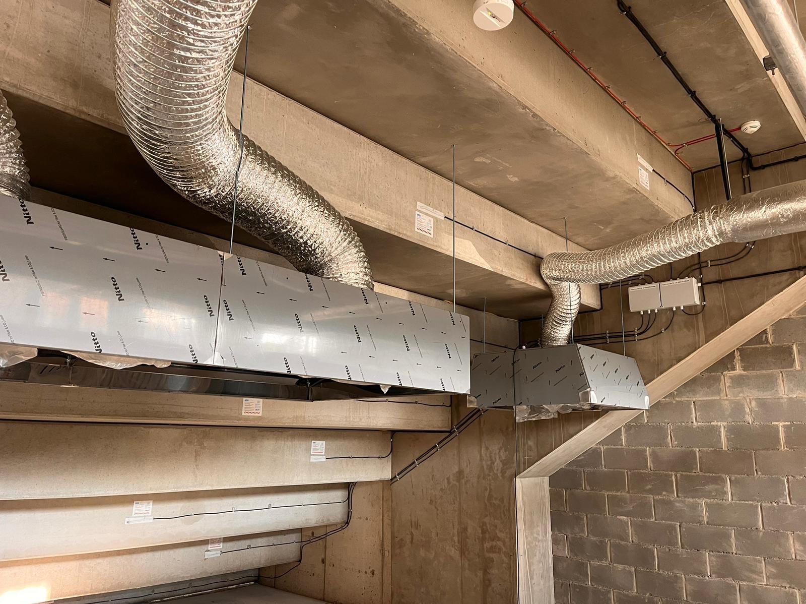 Szare, metalowe kanały wentylacyjne z elastycznymi rurami podwieszone pod betonowym sufitem w surowym wnętrzu budynku. Widoczne zabezpieczenia folią ochronną z logo producenta na elementach kanałów.