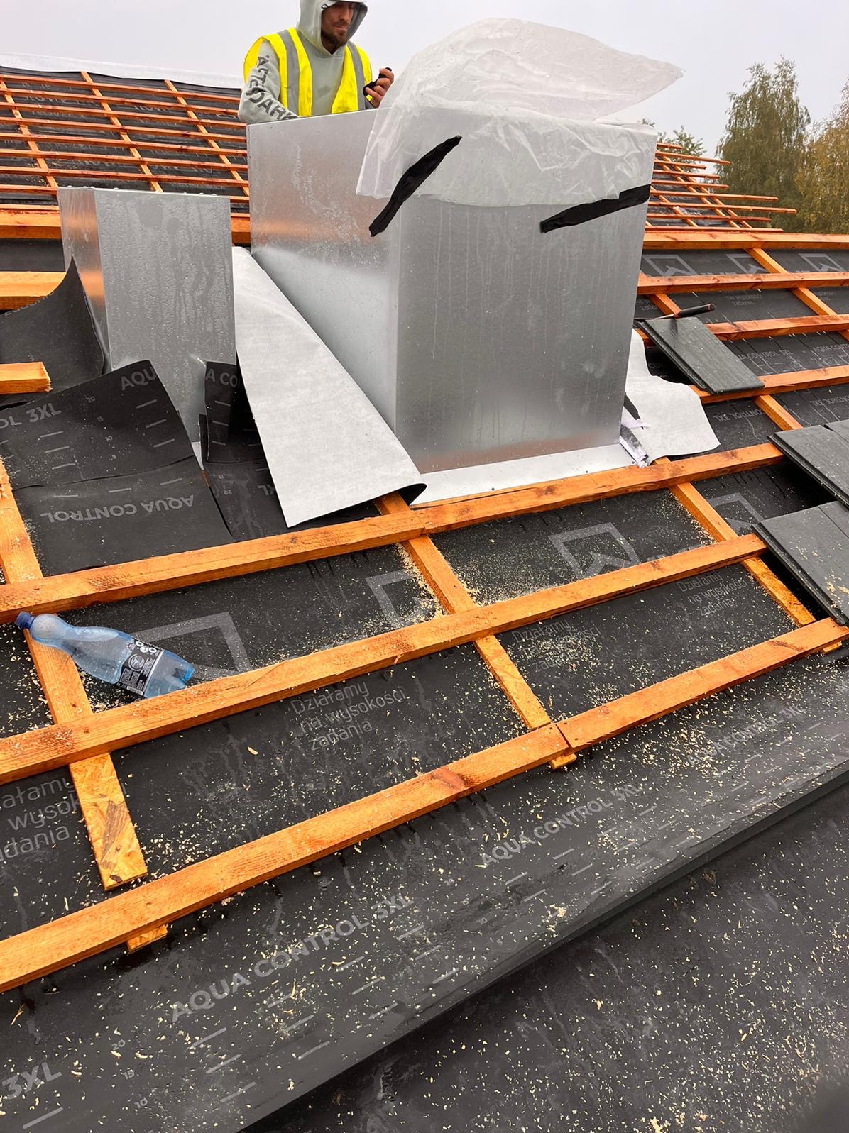 Montaż blaszanej obudowy komina na dachu z drewnianą konstrukcją, widoczna czarna membrana dachowa i częściowo ułożone dachówki, pracownik w odblaskowej kamizelce przytrzymuje element konstrukcji.