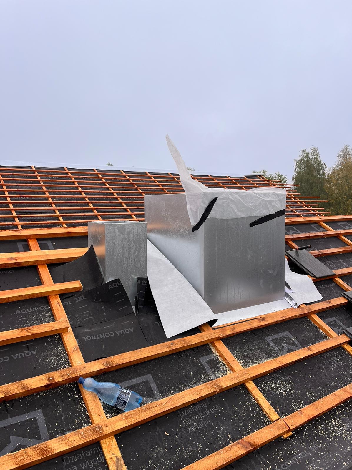 Montaż metalowego komina wentylacyjnego na dachu z drewnianą konstrukcją, zabezpieczonego czarną membraną, widoczna butelka wody i fragmenty posypki dekarskiej.