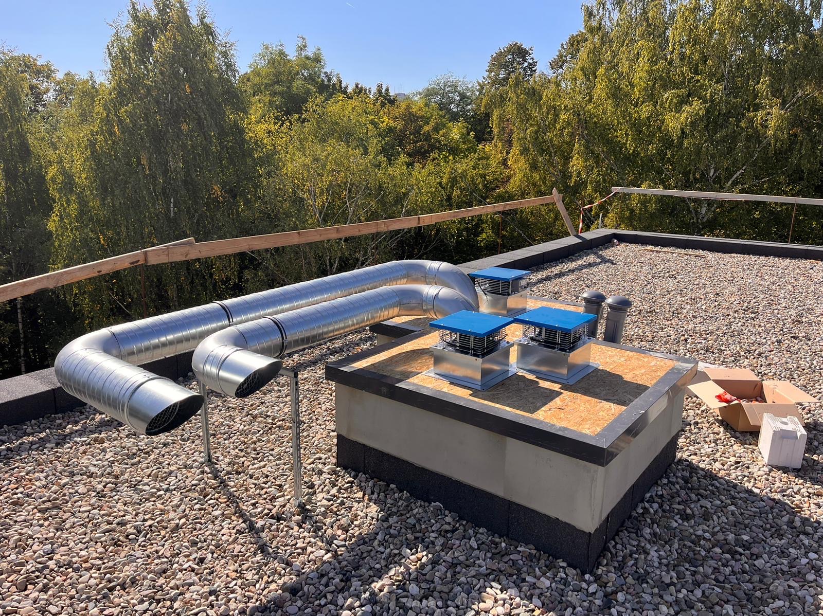 Dwa srebrne, metalowe przewody wentylacyjne na dachu pokrytym żwirem, prowadzące do trzech kominów wentylacyjnych z niebieskimi daszkami. W tle widoczne drzewa.