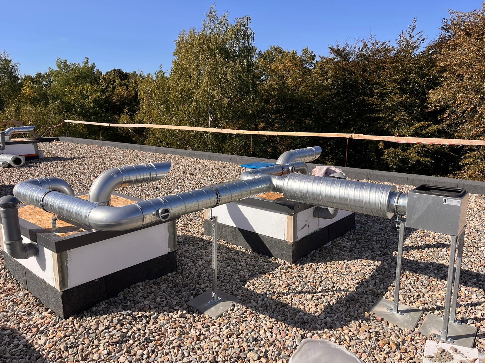 System wentylacyjny z metalowymi rurami spiralnymi na płaskim dachu pokrytym żwirem, z widocznymi elementami mocującymi i drzewami w tle pod błękitnym niebem.