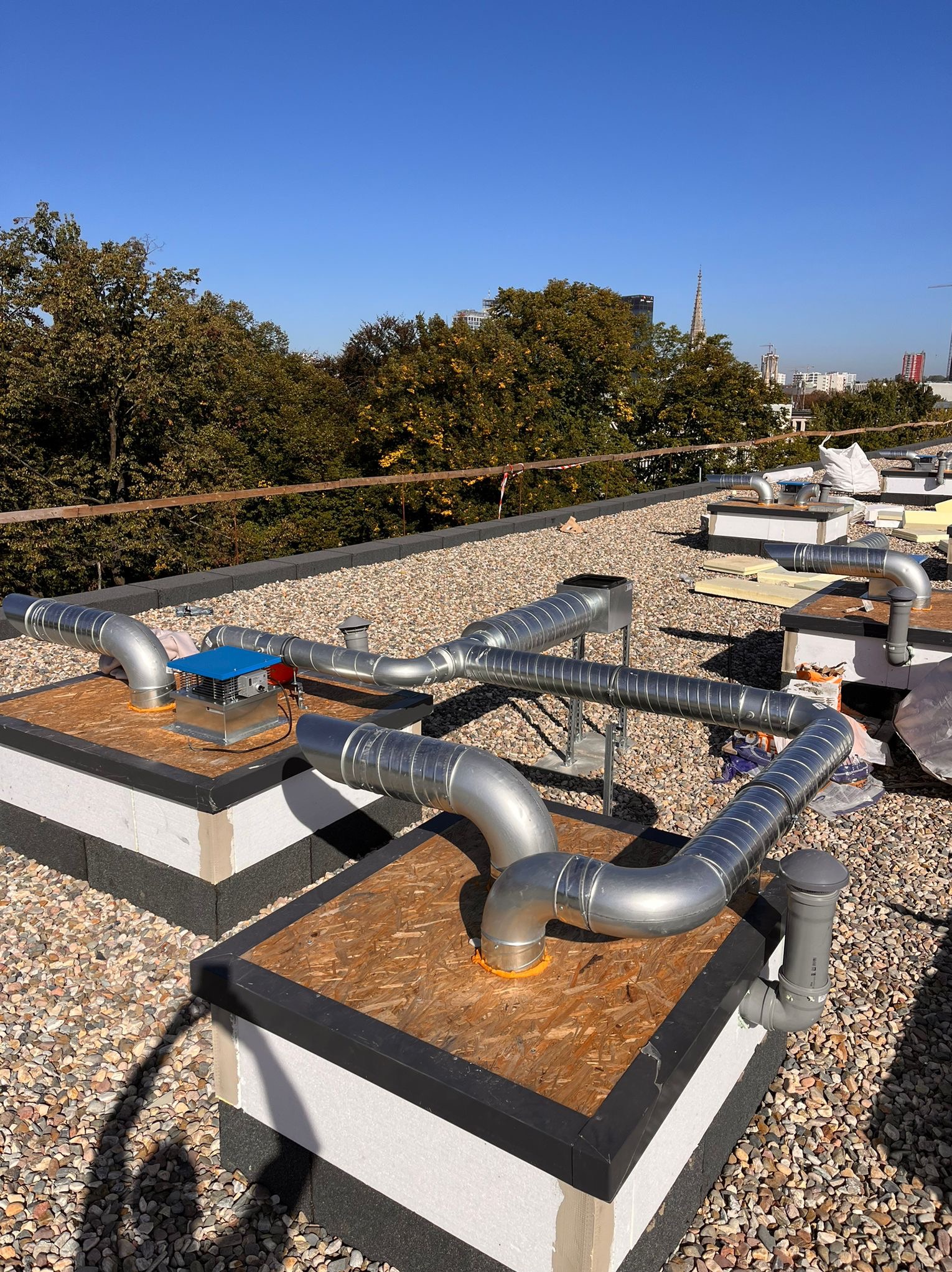 Instalacja wentylacyjna z metalowymi rurami na płaskim dachu pokrytym żwirem, widoczne elementy montażowe i tło miejskie z drzewami i budynkami w oddali.