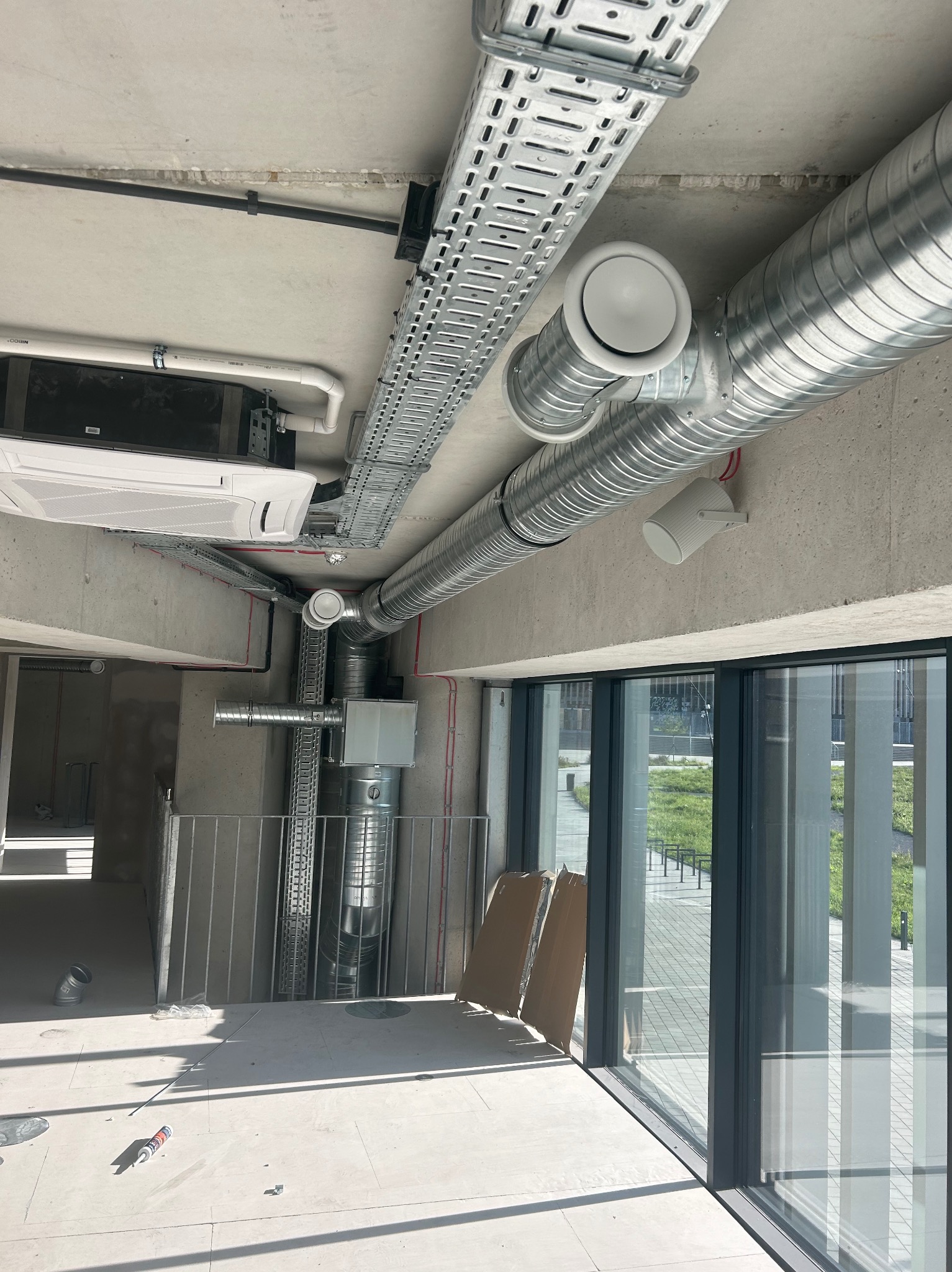 Wnętrze w trakcie instalacji systemu wentylacyjnego: widoczne metalowe rury wentylacyjne pod sufitem, kanały kablowe, klimatyzator kasetonowy, okna z widokiem na zewnątrz, oraz narzędzia na podłodze.