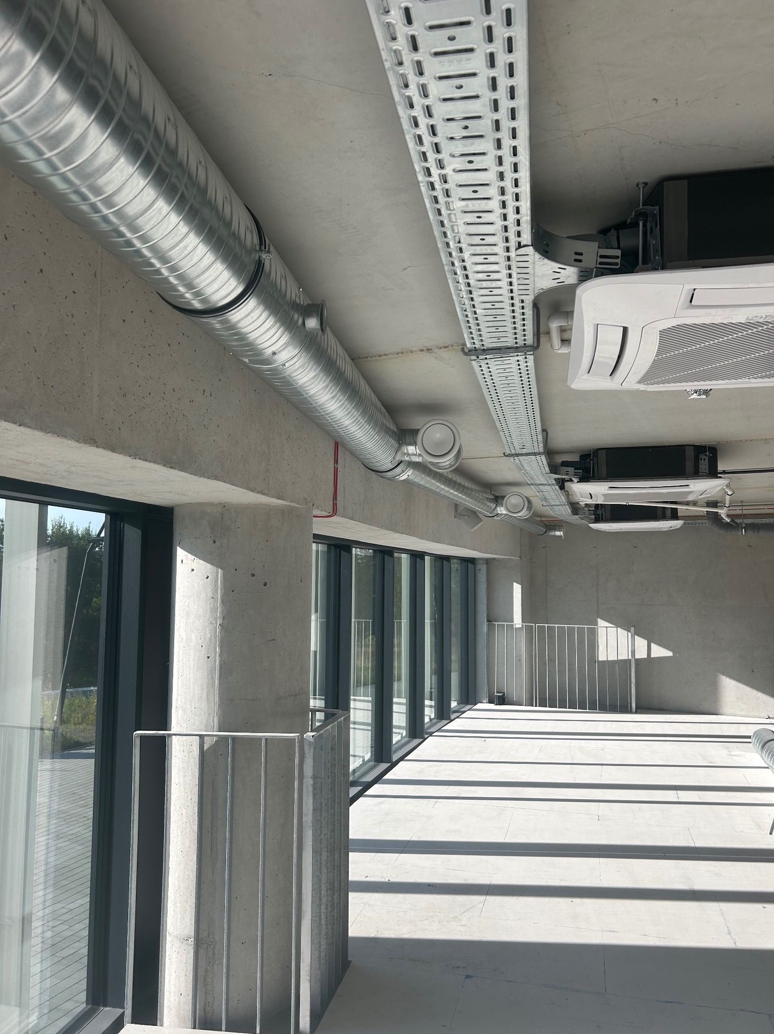 Wnętrze biurowe w surowym stylu z widocznymi elementami instalacji: okrągłe kanały wentylacyjne pod sufitem, koryta kablowe i klimatyzatory kasetonowe.