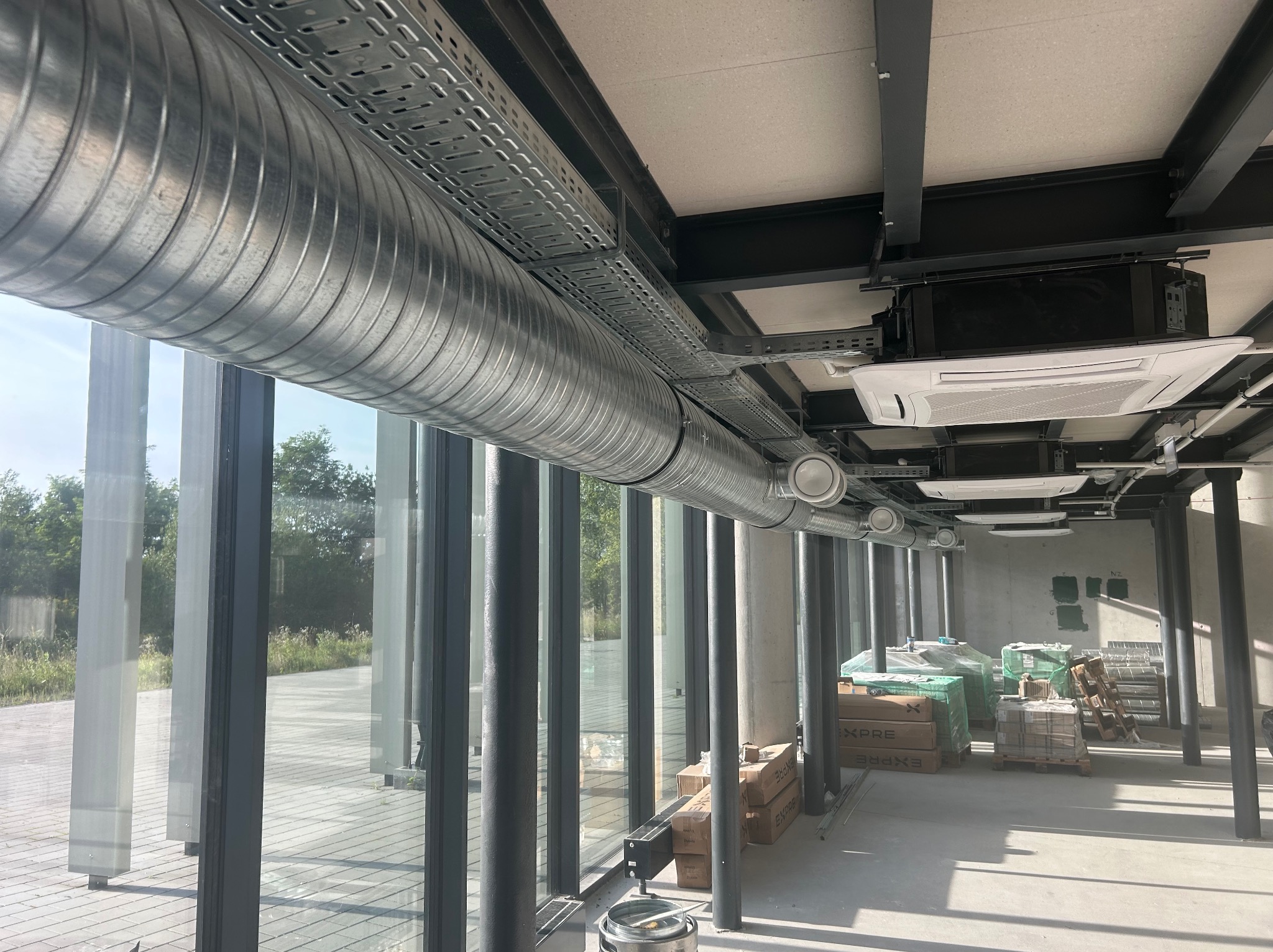 Wnętrze biurowe w trakcie instalacji systemu wentylacji i klimatyzacji, widoczne okrągłe kanały wentylacyjne pod sufitem, kaseta klimatyzacyjna oraz palety z materiałami budowlanymi przy szklanej...