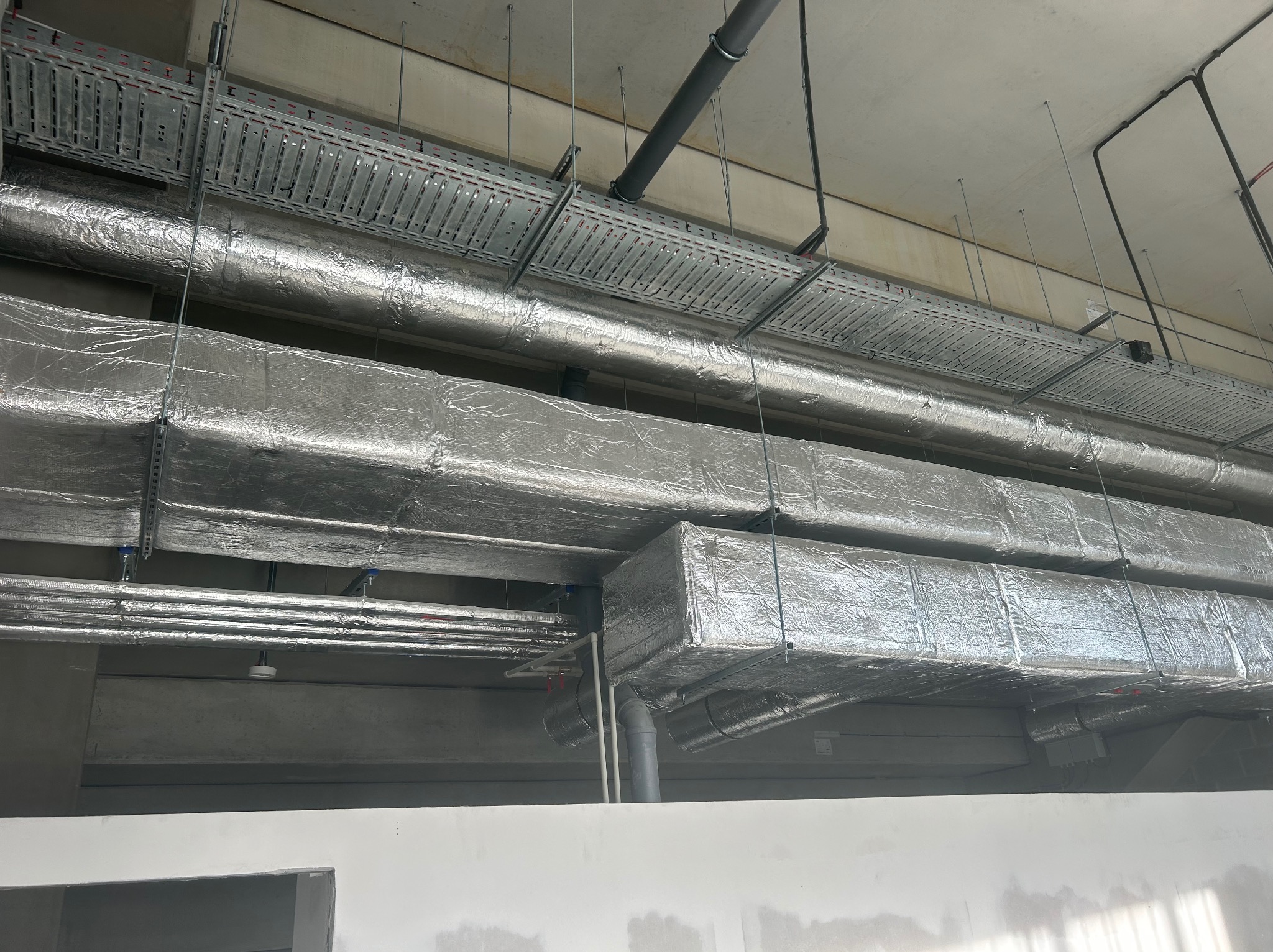 Rury wentylacyjne owinięte folią izolacyjną, zawieszone pod sufitem z widocznymi elementami konstrukcyjnymi i metalowymi korytami kablowymi.