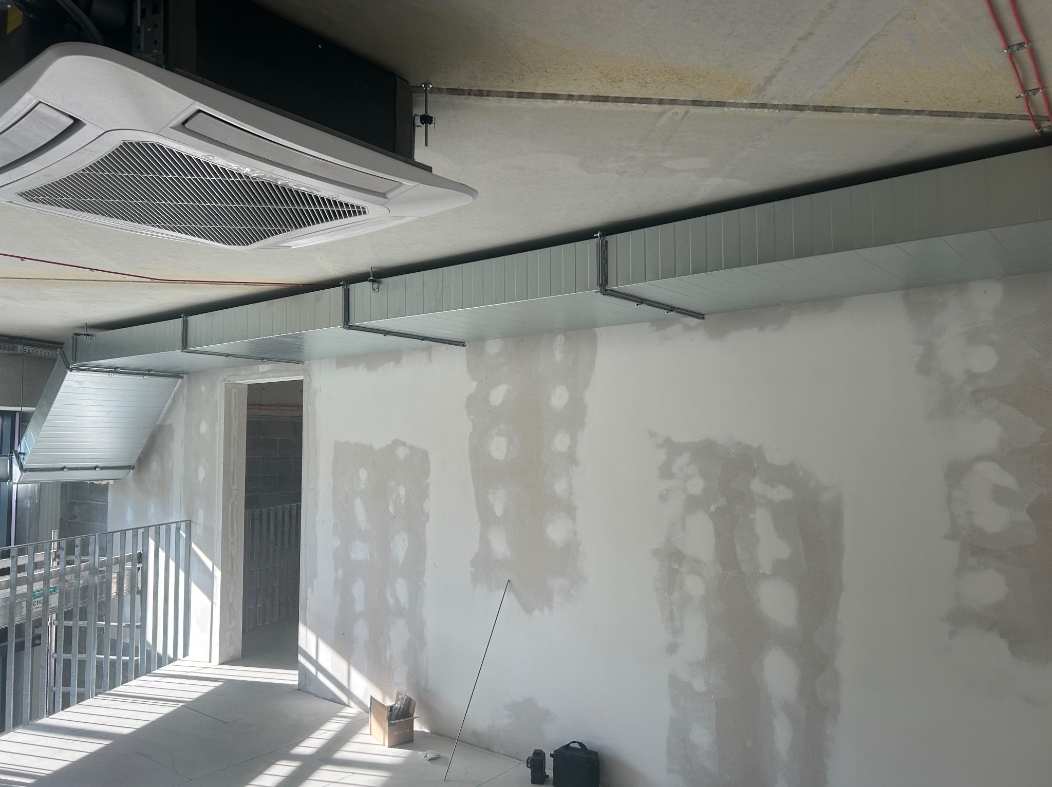 Wnętrze w trakcie wykańczania z zamontowaną kasetą klimatyzacyjną sufitową i kanałami wentylacyjnymi pod stropem. Ściany z widocznymi śladami szpachlowania, podłoga z resztkami gruzu.