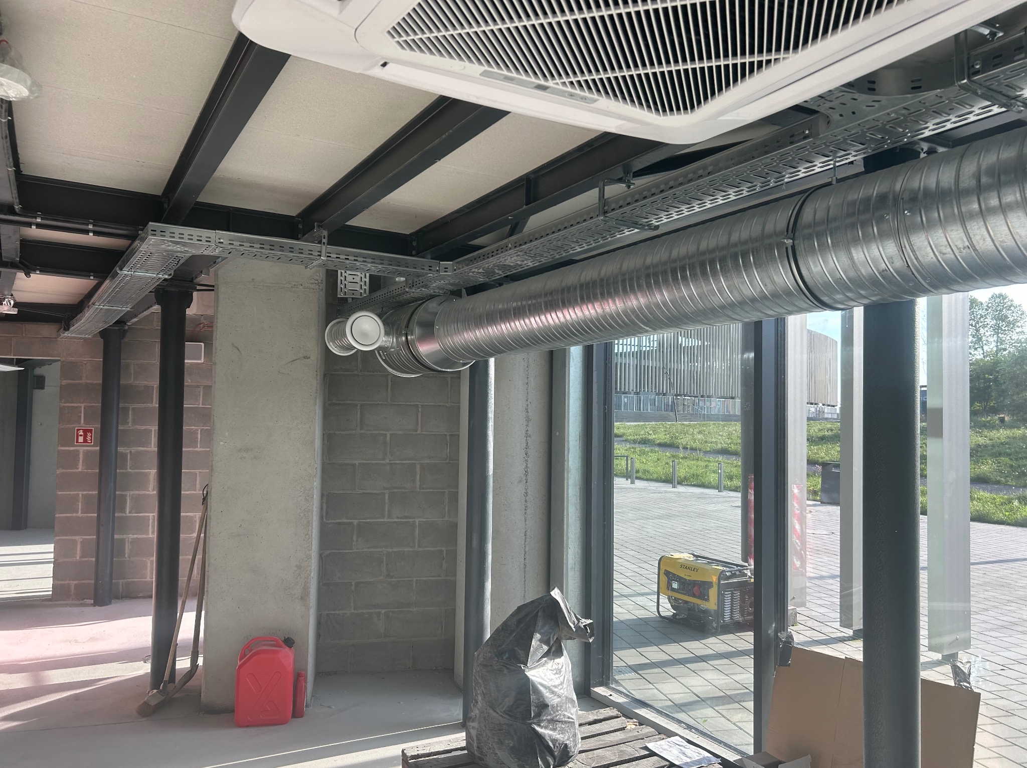 Instalacja wentylacji: rura wentylacyjna z blachy falistej, zawieszona pod sufitem na metalowych konstrukcjach, widoczny klimatyzator kasetonowy, betonowe ściany i słupy, na zewnątrz widoczny...