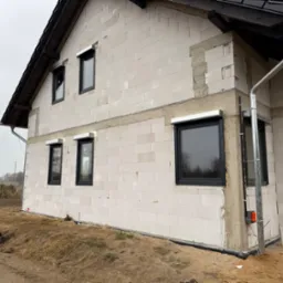 Surowy stan domu w trakcie budowy z zamontowanymi oknami w czarnych ramach i roletami zewnętrznymi, widoczne bloczki z betonu komórkowego i fragment systemu rynnowego.