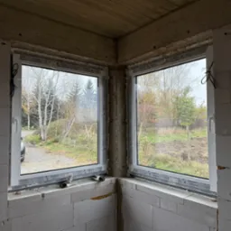 Dwa białe okna narożne w surowym stanie budynku, widoczne uszczelnienia i pianka montażowa, za oknami widok na jesienne drzewa i podwórko.