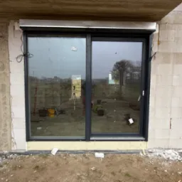 Nowe, czarne, przesuwne okno PCV z zamontowaną roletą zewnętrzną w surowym stanie, widoczne narzędzia i niedokończone wykończenie muru.