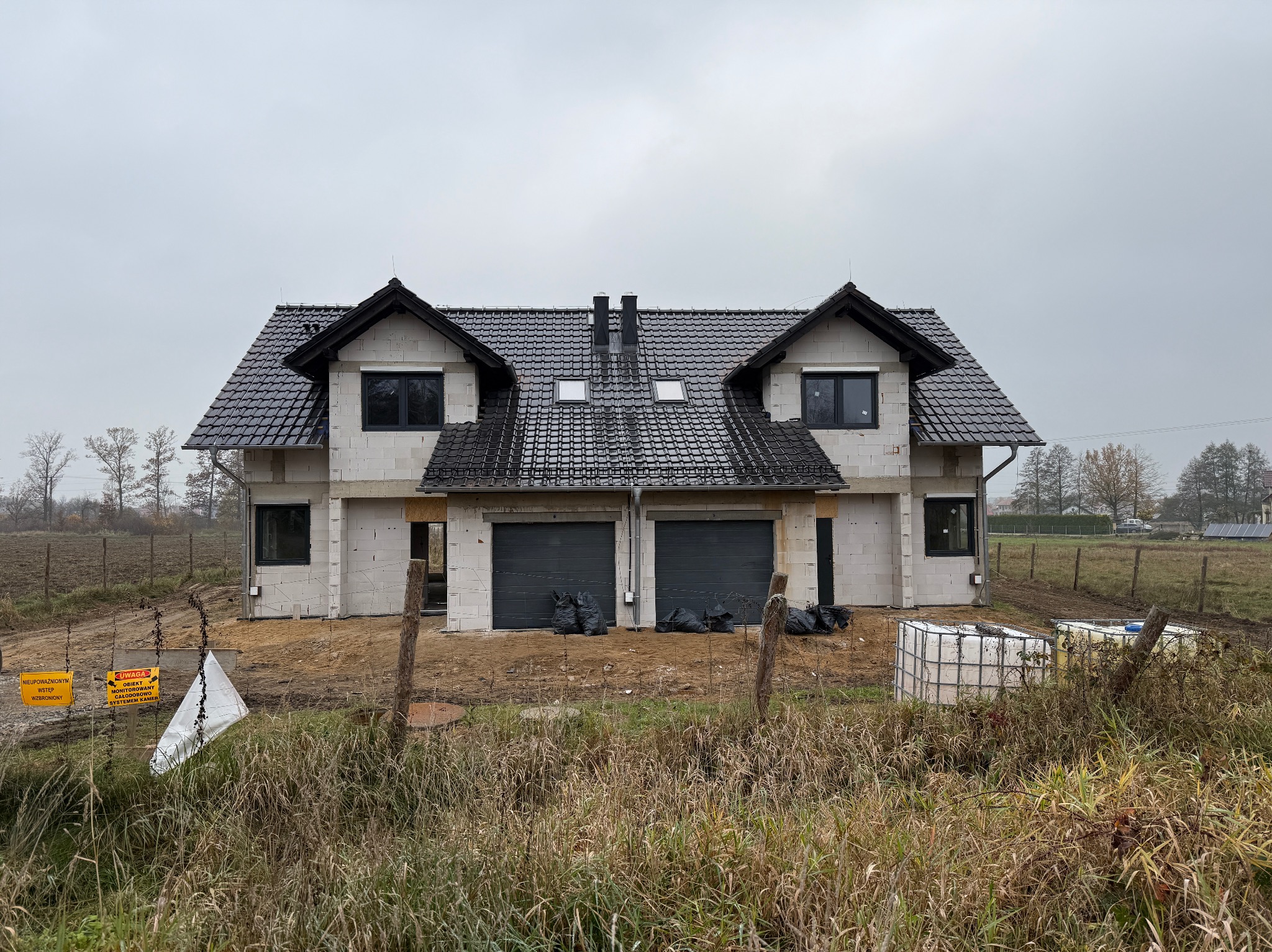 Surowy stan bliźniaka z szarego bloczka, czarne ramy okienne i garażowe drzwi, czarna dachówka, teren budowy zabezpieczony płotem z ostrzeżeniami, pochmurne niebo.