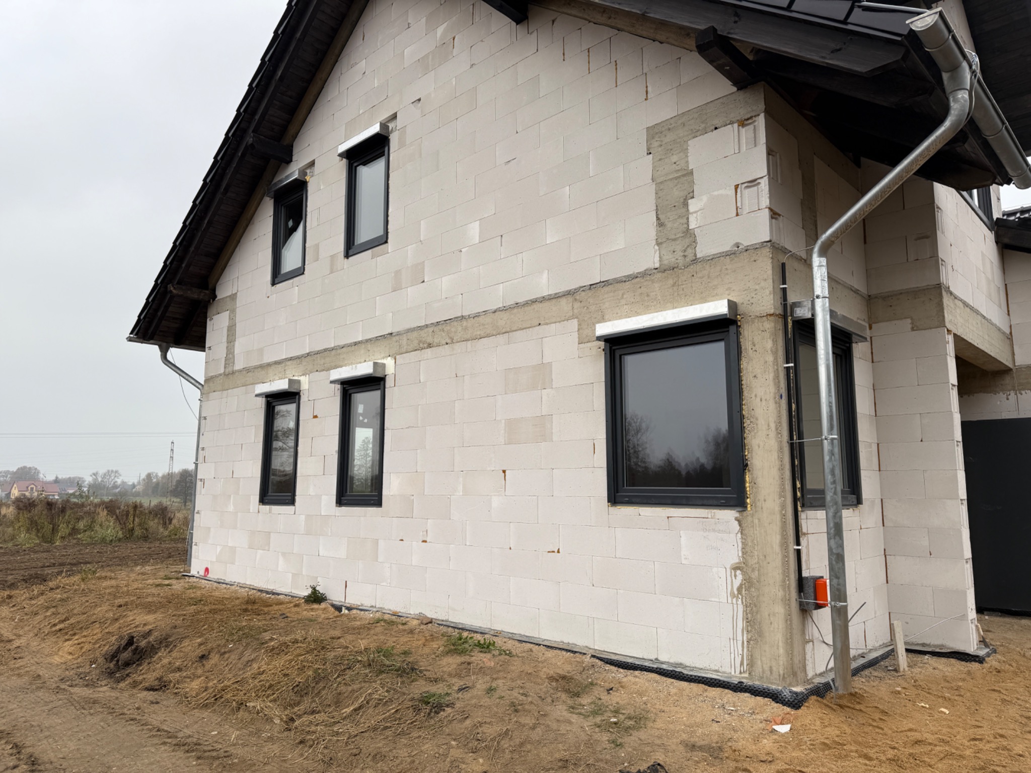 Surowy stan domu w trakcie budowy z zamontowanymi oknami w czarnych ramach i roletami zewnętrznymi, widoczne bloczki z betonu komórkowego i fragment systemu rynnowego.