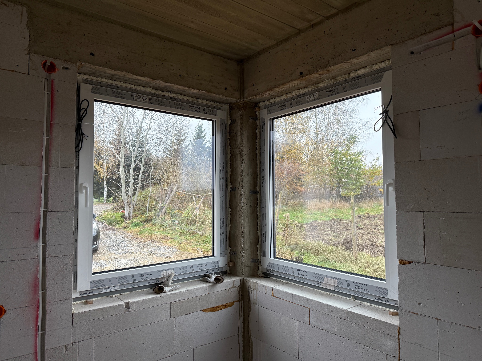 Dwa białe okna narożne w surowym stanie budynku, widoczne uszczelnienia i pianka montażowa, za oknami widok na jesienne drzewa i podwórko.