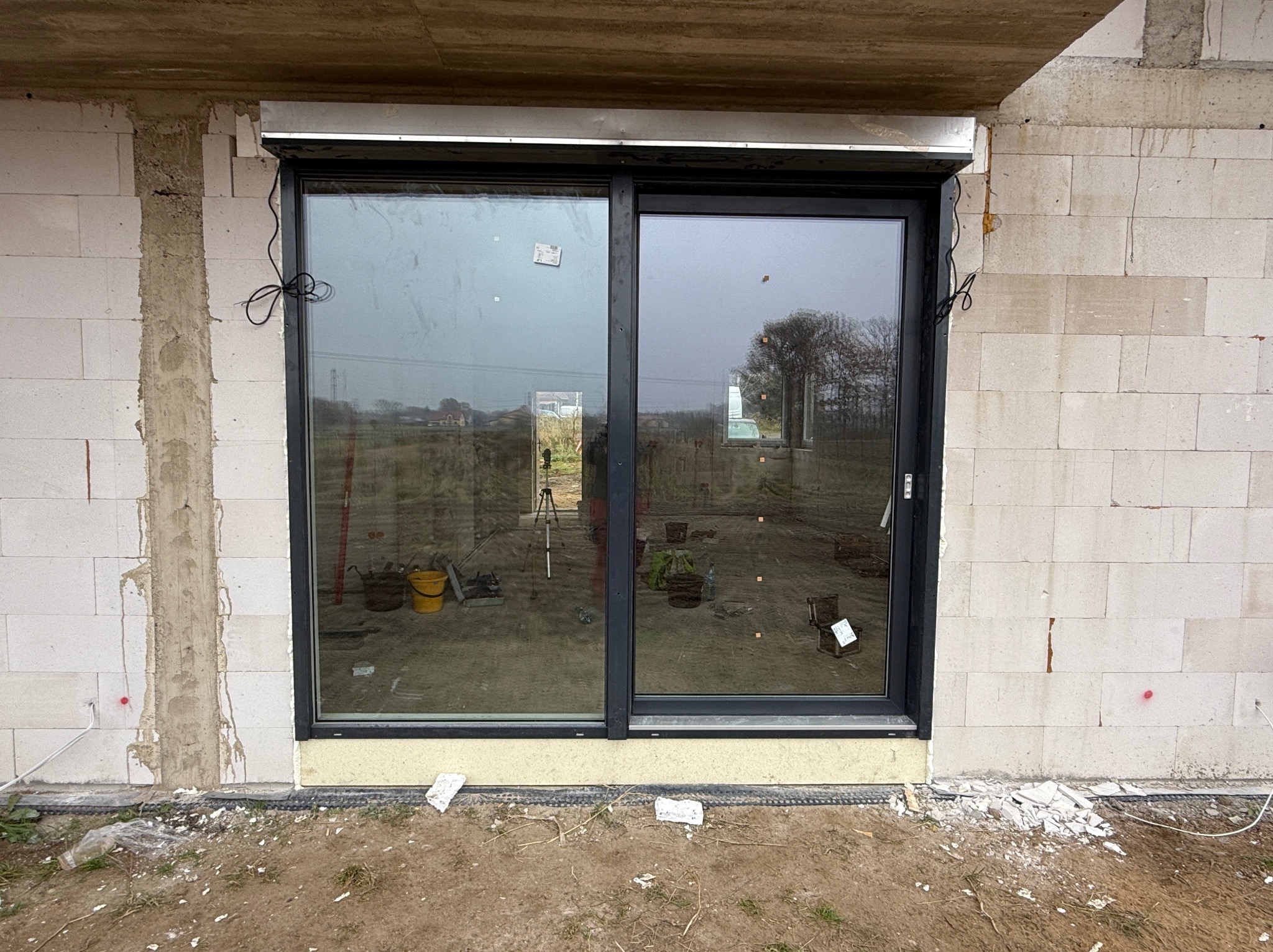 Nowe, czarne, przesuwne okno PCV z zamontowaną roletą zewnętrzną w surowym stanie, widoczne narzędzia i niedokończone wykończenie muru.