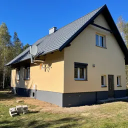 Elewacja domu jednorodzinnego w odcieniach beżu i szarości, z widoczną anteną satelitarną i fragmentem ogrodu. Dom otoczony drzewami, błękitne niebo w tle.