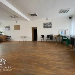 Karol Ebertowski Usługi Ogólnobudowlane - Sala po remoncie z drewnianą podłogą, białymi ścianami i sufitami. Widoczne krzesła, stoły i oświetlenie. Na ścianach wiszą obrazy. Charakterystyczny wzór parkietu.