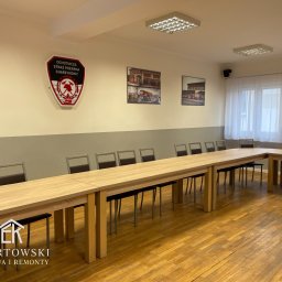 Karol Ebertowski Usługi Ogólnobudowlane - Jasne, odnowione wnętrze sali konferencyjnej z długim stołem i krzesłami. Na ścianie logo OSP i zdjęcia. Parkiet po renowacji. Widoczny logotyp firmy remontowej.