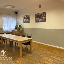 Karol Ebertowski Usługi Ogólnobudowlane - Jasne biuro po odświeżeniu: drewniana podłoga, stół konferencyjny, szare ściany do połowy wysokości, zdjęcia w ramkach na górze. Widok na okno z białymi firankami.