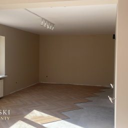 Karol Ebertowski Usługi Ogólnobudowlane - Układanie parkietu w jodełkę w pokoju z beżowymi ścianami i białym oknem. Część podłogi jeszcze nieukończona, widoczny surowy beton. Na ścianie widoczne oświetlenie.