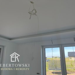 Karol Ebertowski Usługi Ogólnobudowlane - Białe wnętrze po remoncie z widocznymi przewodami elektrycznymi, listwami sufitowymi i oknami. Na ścianie logo firmy budowlanej.