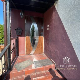 Karol Ebertowski Usługi Ogólnobudowlane - Wejście do domu z nowoczesnymi drzwiami z owalnym przeszkleniem, brązową elewacją i terakotą na schodach. Widoczna skrzynka pocztowa i cień osoby.