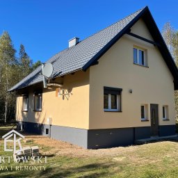 Karol Ebertowski Usługi Ogólnobudowlane - Żółty dom jednorodzinny z szarym dachem i podmurówką, widziany z boku w słoneczny dzień. Widoczne okna z roletami, antena satelitarna i fragment ogrodu.